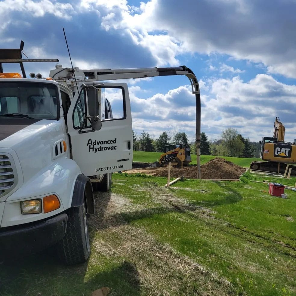 Advanced Hydrovac truck vacuuming soil at grassy construction site with excavators and cloudy blue sky