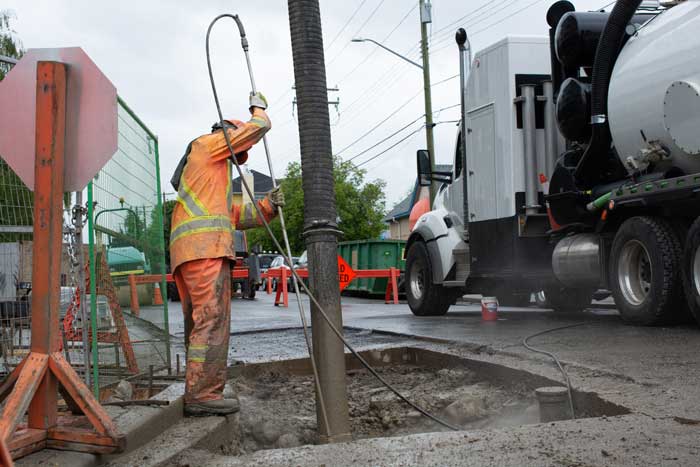 Construction worker in orange hi-vis gear using vacuum hose to clean a manhole beside a utility truck on street