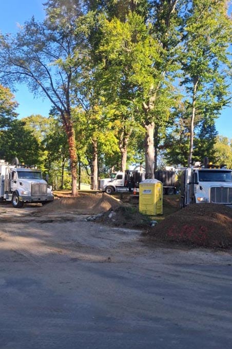 Construction site with three dump trucks, portable toilet, dirt piles and tall trees under blue sky