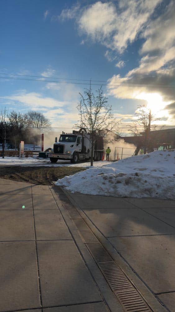 Work truck and crew member in high-visibility jacket near steaming manhole in snowy urban sidewalk at sunset.