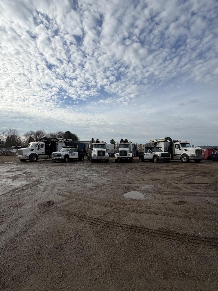 Row of white industrial work trucks parked on a muddy lot beneath an expansive cloud-filled sky