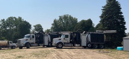 Two large industrial vacuum excavation trucks parked on a dirt lot with trees in the background