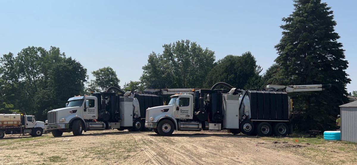 Two large industrial vacuum excavation trucks parked on a dirt lot with trees in the background