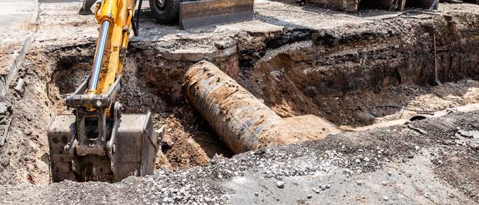 Backhoe excavating a construction trench exposing a large rusted underground pipeline.