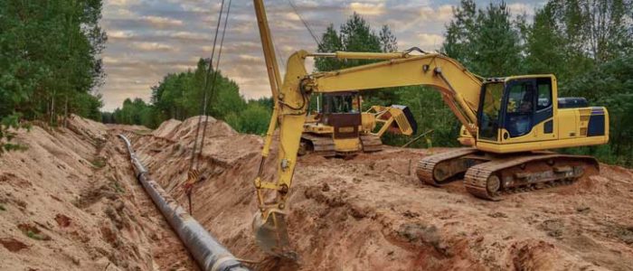 Yellow excavator installing large steel pipeline in deep trench at rural construction site under a cloudy sky