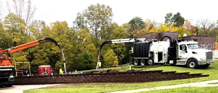 Industrial vacuum trucks with articulated booms and workers removing debris by a brick retaining wall amid autumn trees.